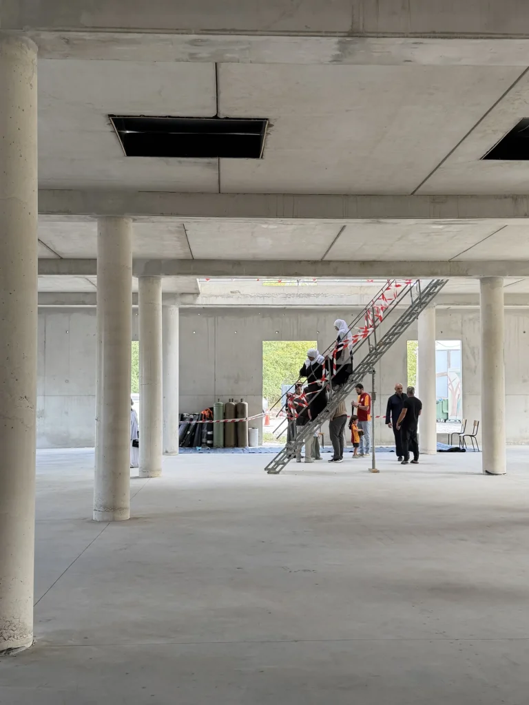 Des visiteurs au pied d’un escalier de chantier, dans une grande salle aux colonnes de béton.